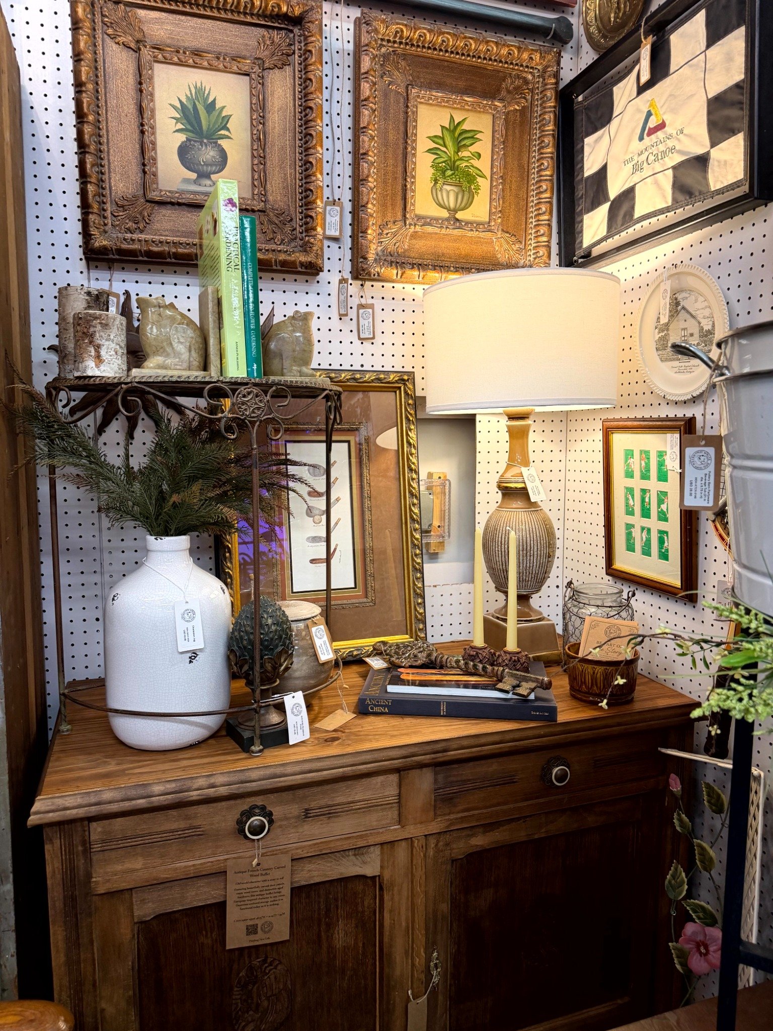 A curated arrangement of botanical artwork, a glowing table lamp, ceramics, and books on an antique wooden cabinet.